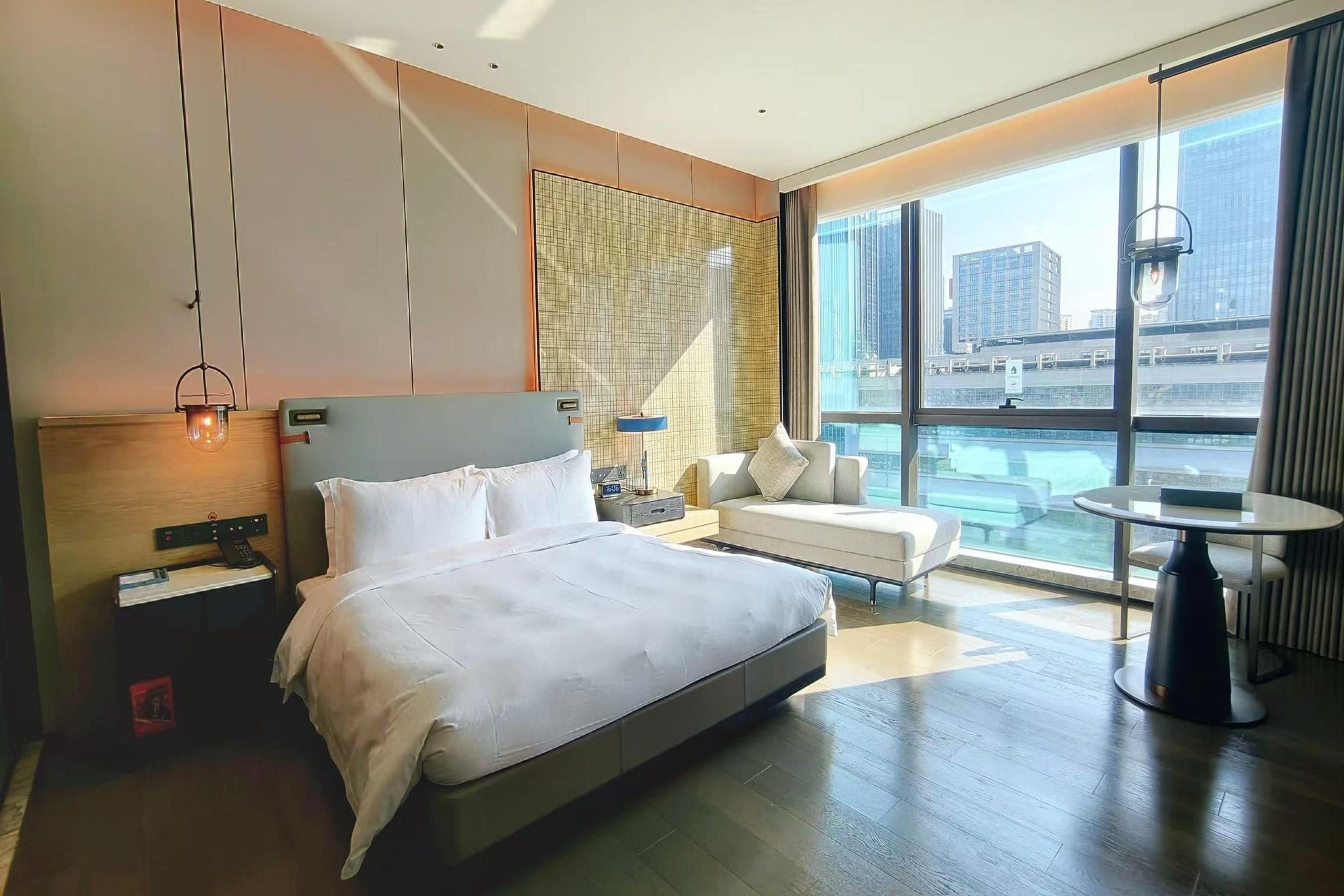 A modern hotel room with a large bed, a seating area with a sofa and armchairs, a small table, and a city view through large windows.