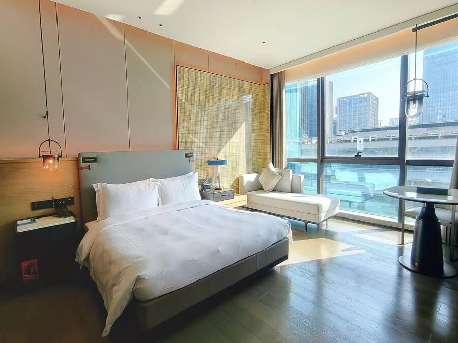 Habitación de hotel moderna con una cama grande, zona de estar con sofá y sillones, una mesita y vistas a la ciudad a través de amplios ventanales.
