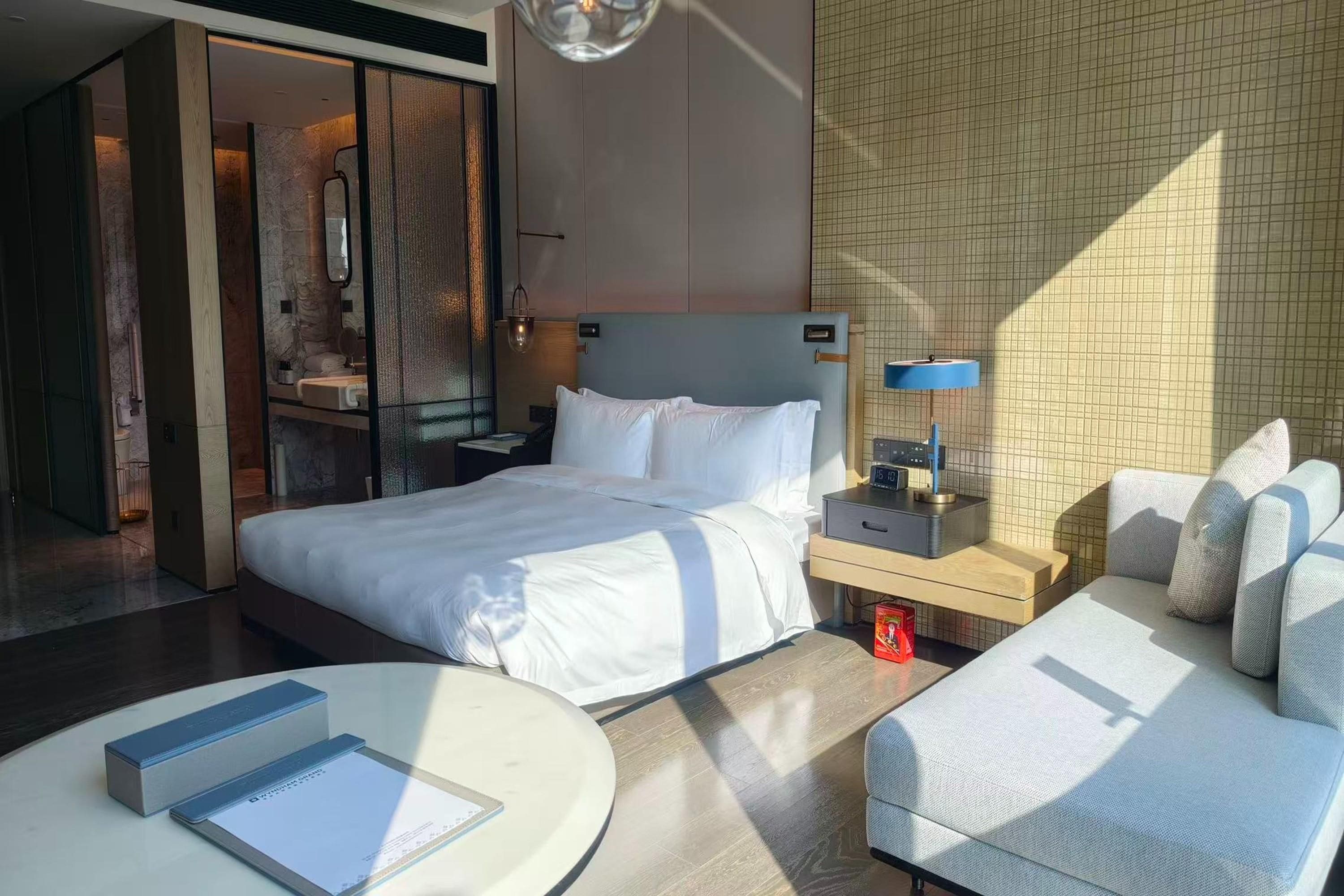 A modern hotel room with a large bed, bedside table, and a seating area with a sofa and armchairs.