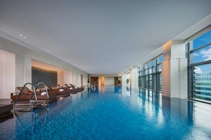 An indoor swimming pool with clear blue water, lounge chairs, and a view of the city through large windows.