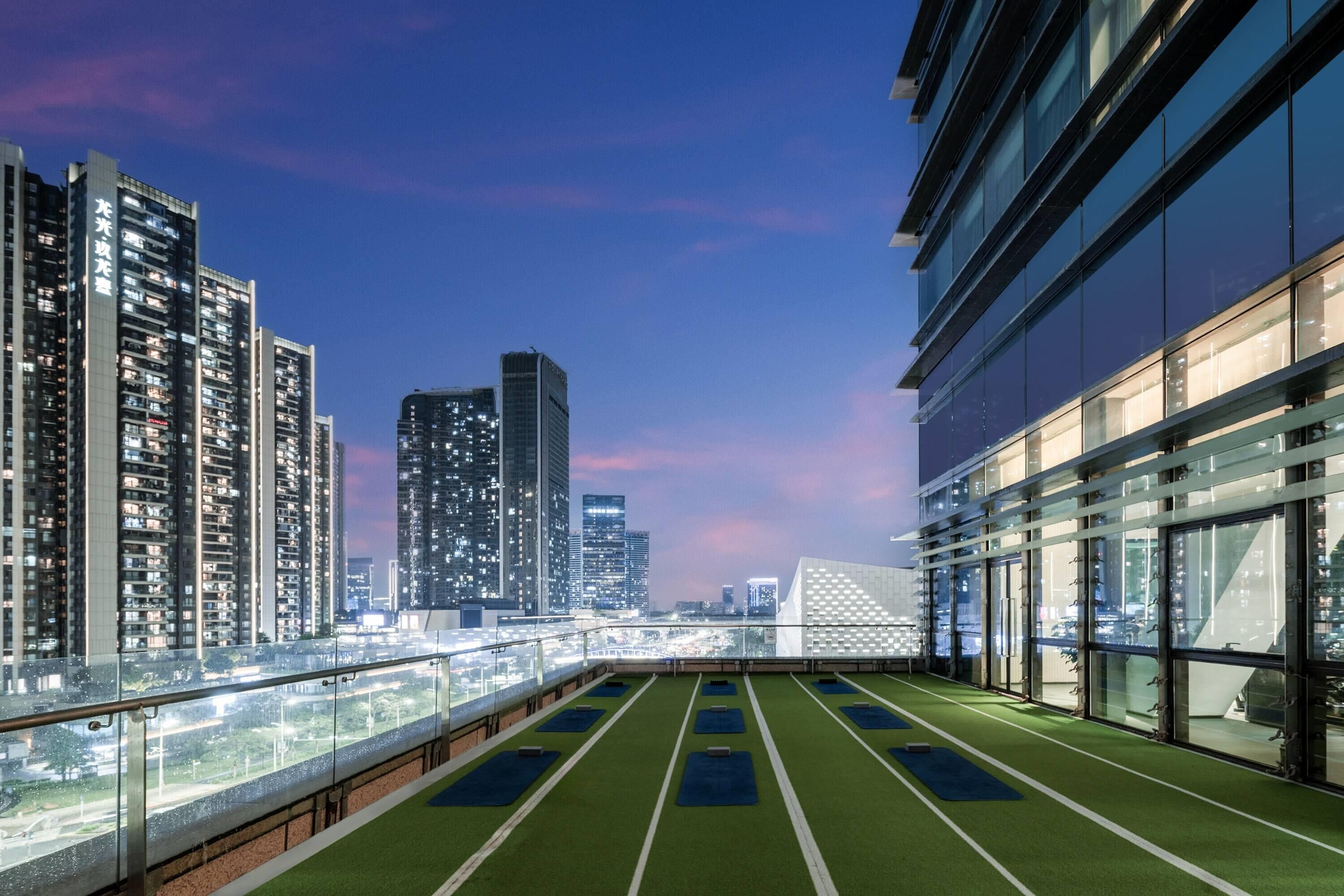 A rooftop with green artificial turf and exercise mats, overlooking a cityscape with high-rise buildings and a distinctive white structure.