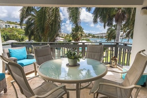 A round glass table with wicker chairs and a blue cushion, set on a balcony with a view of a marina and palm trees. - Tropical Lotus- Cozy 3 bedroom, 3 bath with an ocean view, pool, gym, & tennis (St Thomas)