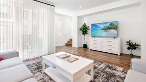A modern living room with a white sofa, a coffee table with magazines, a TV on a white cabinet, and a large window with sheer curtains. - Central Location Cozy Private Bedroom w Bathroom (Irvine)