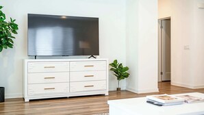 A modern living room with a flat-screen TV on a white cabinet, a coffee table with magazines, and a potted plant. - Central Location Cozy Private Bedroom w Bathroom (Irvine)