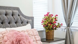 A bedroom with a tufted headboard, a pink floral pillow, a bedside table with a woven basket of flowers, and a window with blinds. - Central Location Cozy Private Bedroom w Bathroom (Irvine)