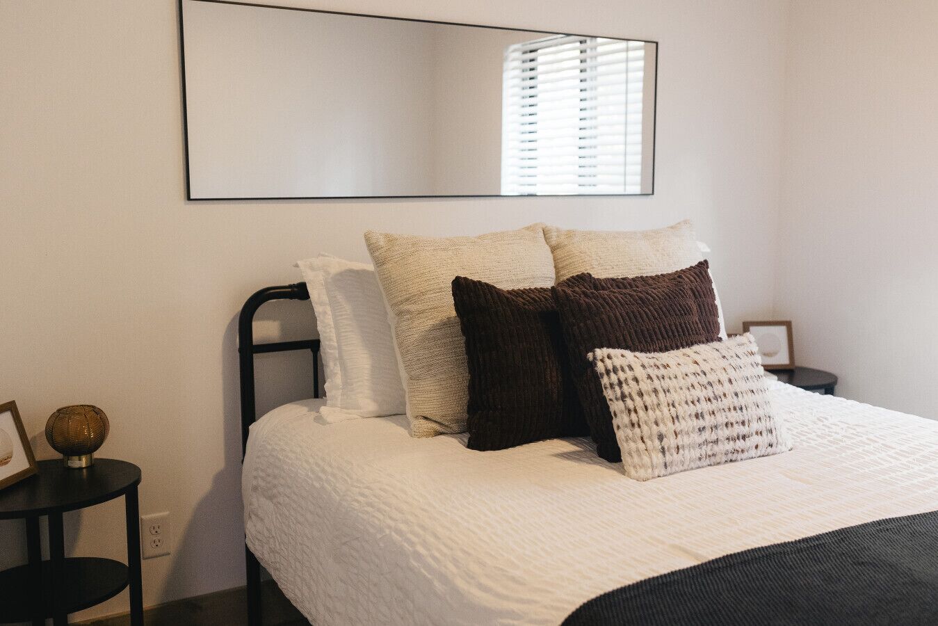 A neatly made bed with a white duvet, multiple pillows, and a black headboard. There is a round table with a decorative item on it, and a framed picture on the wall.