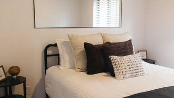 A neatly made bed with a white duvet, multiple pillows, and a black headboard. There is a round table with a decorative item on it, and a framed picture on the wall.