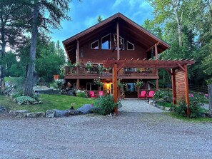 Une cabane en rondins à deux étages, dotée d’un balcon, entourée d’une allée de gravier et d’une végétation luxuriante.
