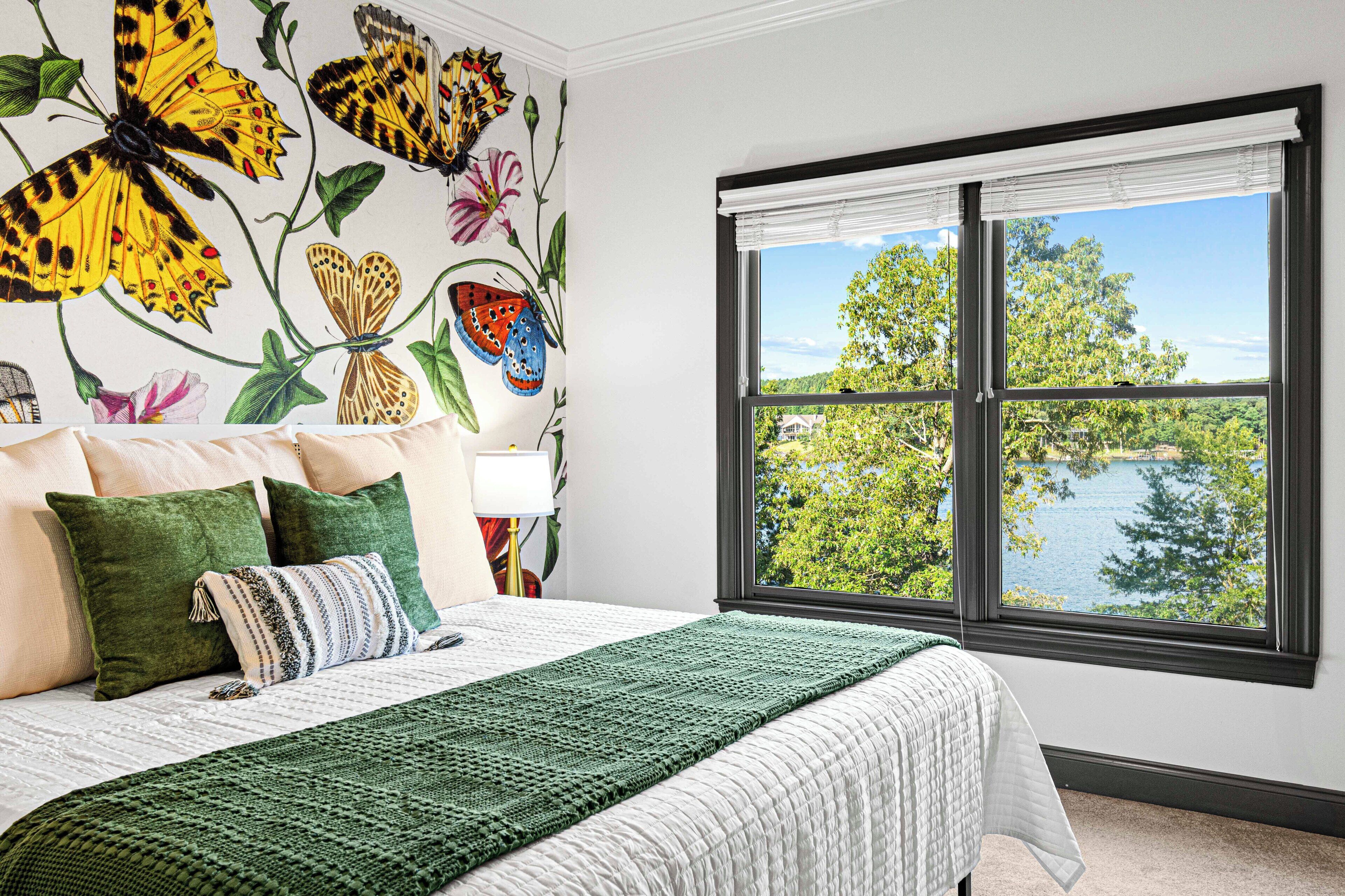 A bedroom with a large bed, green and white bedding, a view of a lake through a window, and a wall mural of butterflies and flowers.