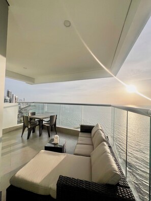 Un balcón moderno con barandilla de vidrio, un sofá, una mesa de comedor con sillas y vistas al horizonte urbano y al agua.