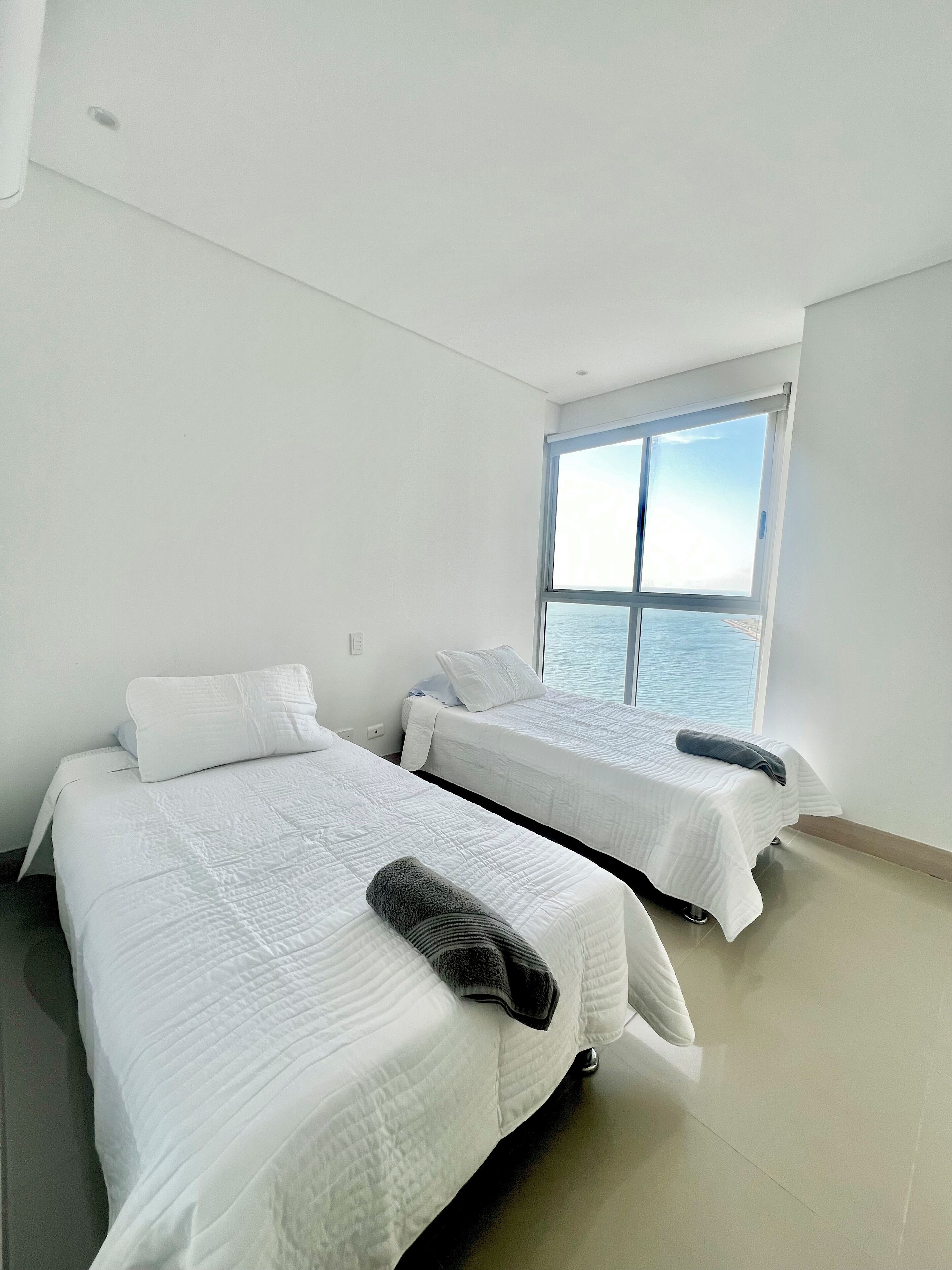 A hotel room with two beds, a large window with a view of the sea, and a tiled floor.