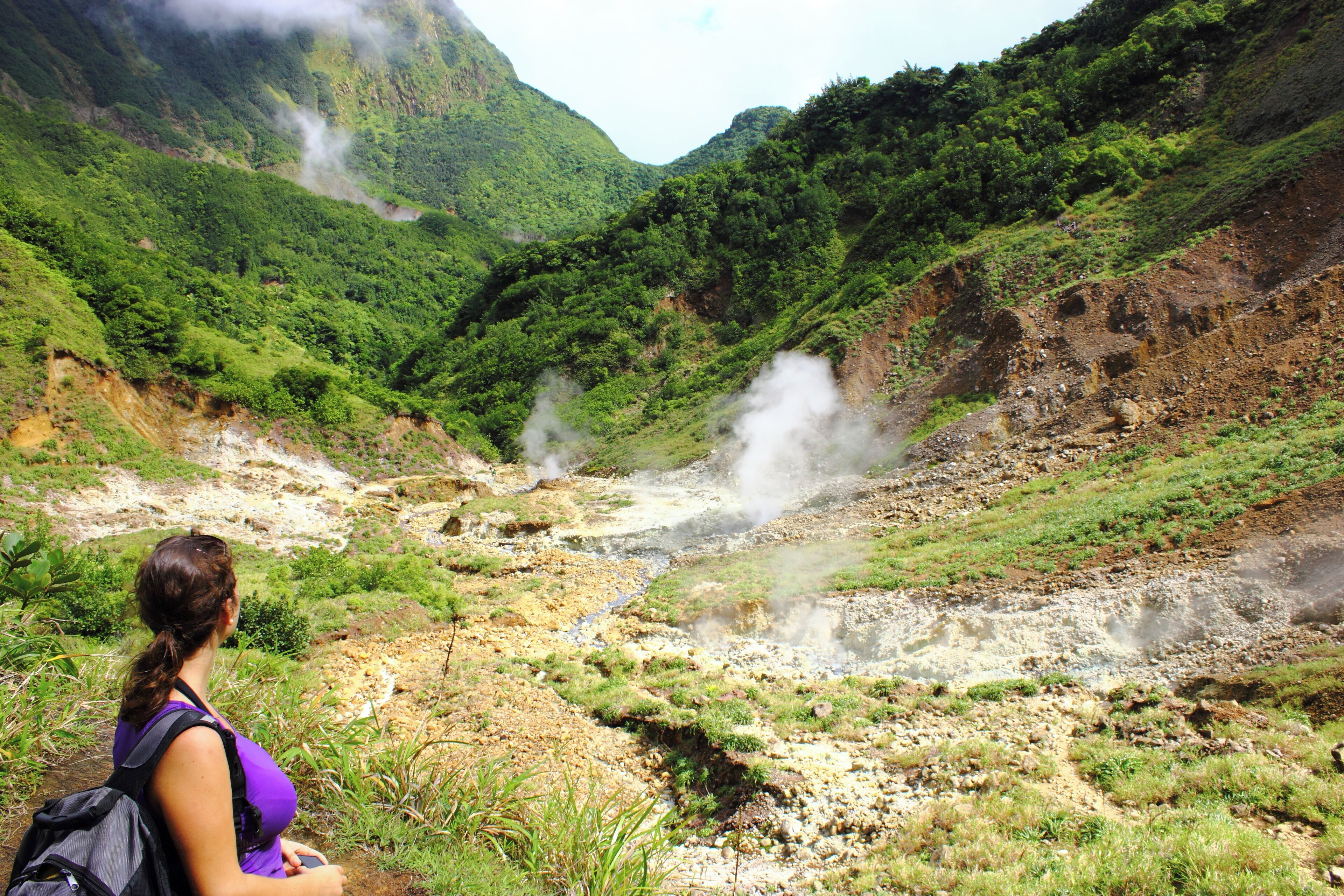 A woman standing on a path overlooking a valley with a steam vent, surrounded by lush greenery and hills.