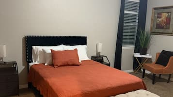 Un dormitorio con cama, banco, ventilador de techo, mesita de noche, lámpara, planta en maceta y una silla.
