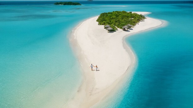 An aerial view of a secluded beach with a small island, clear turquoise waters, and a couple walking along the shore. - Nest by Hawks Hotels Kamadhoo Baa Atoll (Kamadhoo)
