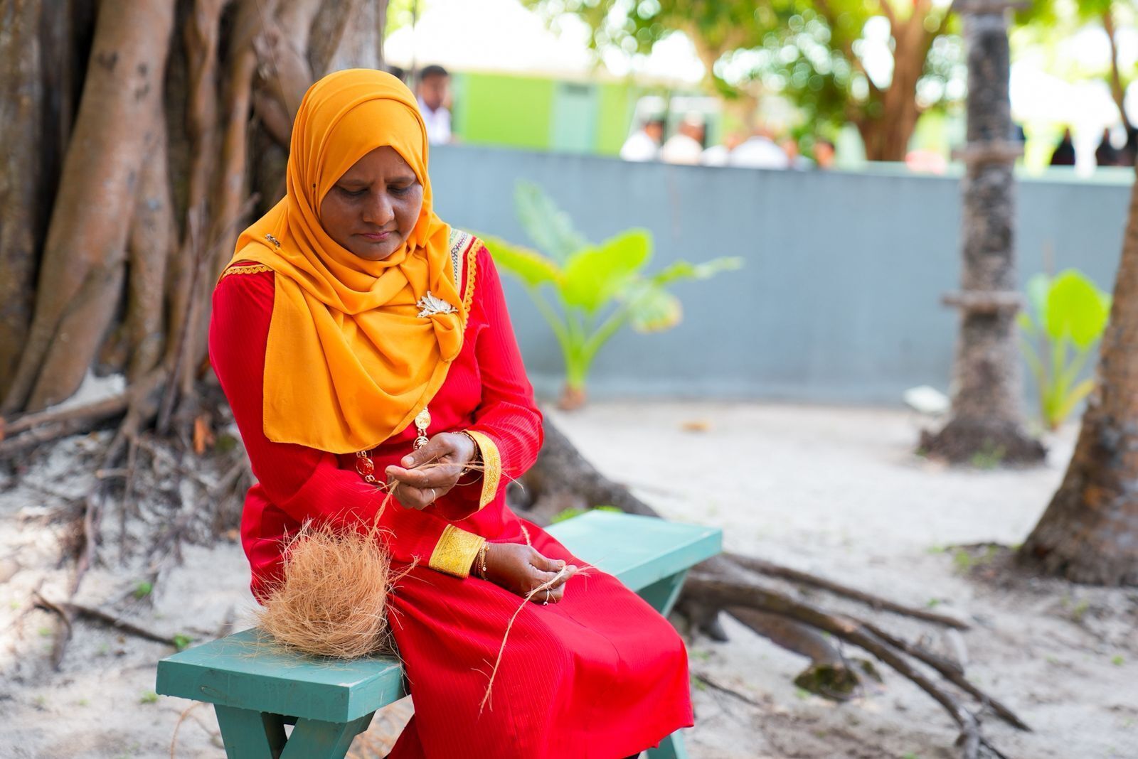 A person sitting on a bench outdoors, wearing a red outfit and a yellow headscarf.