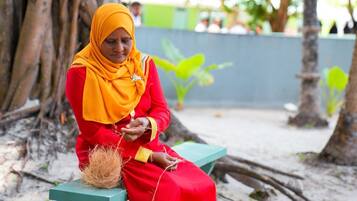 A person sitting on a bench outdoors, wearing a red outfit and a yellow headscarf.
