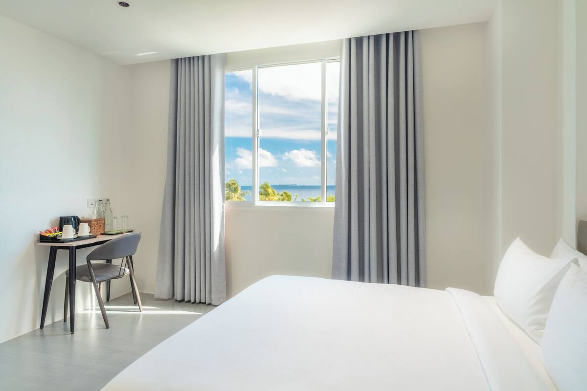 Quarto de hotel com uma cama grande, uma escrivaninha com cadeira e vista para o mar através de uma janela com cortinas.