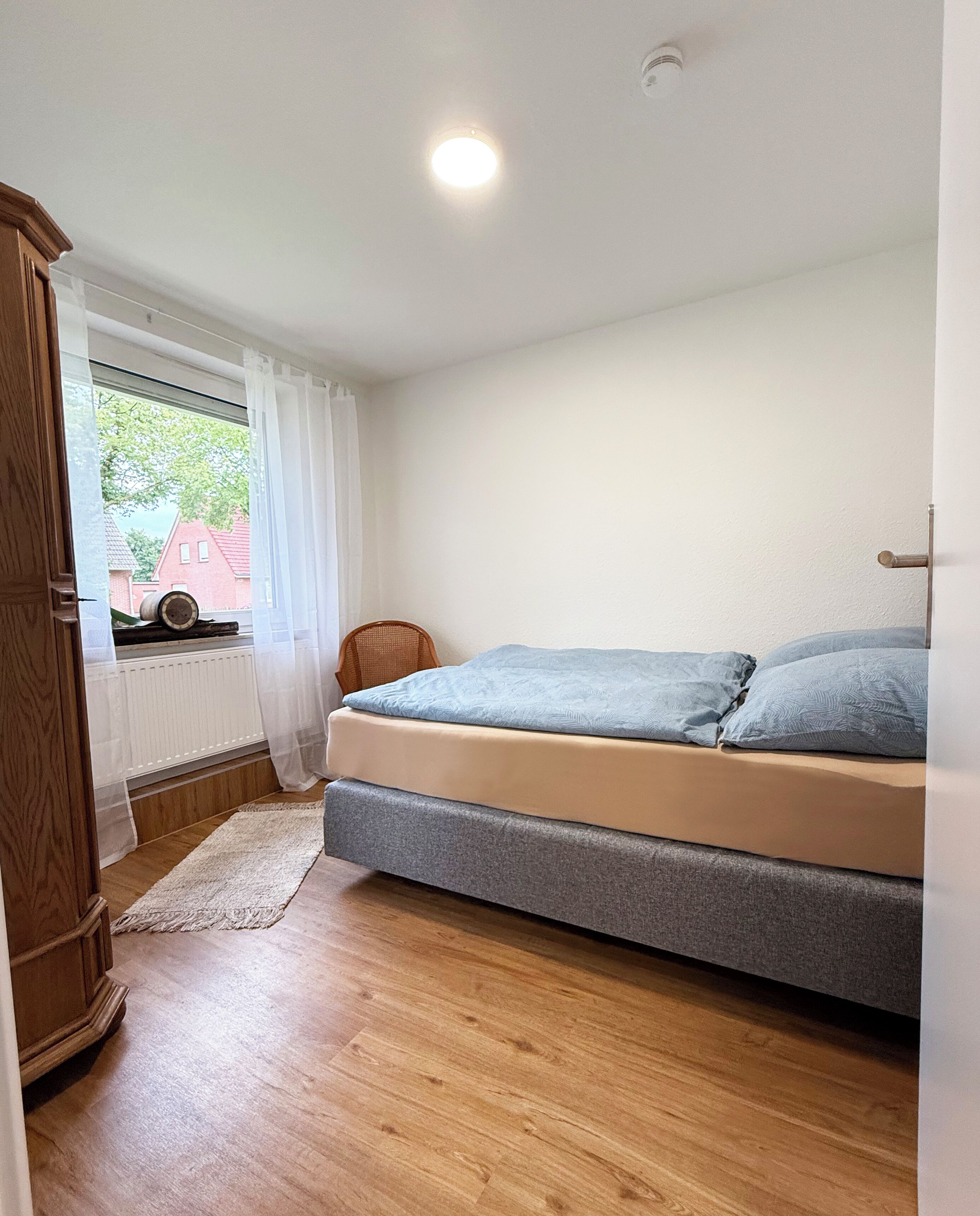 A bedroom with a bed, a wooden wardrobe, a window with curtains, and a view of a red building outside.