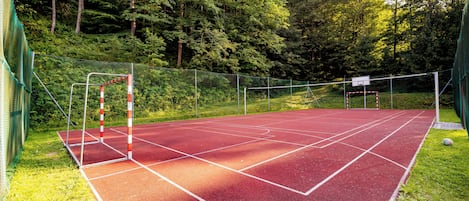 Cancha de tenis exterior roja con una red, rodeada por una valla verde y árboles.