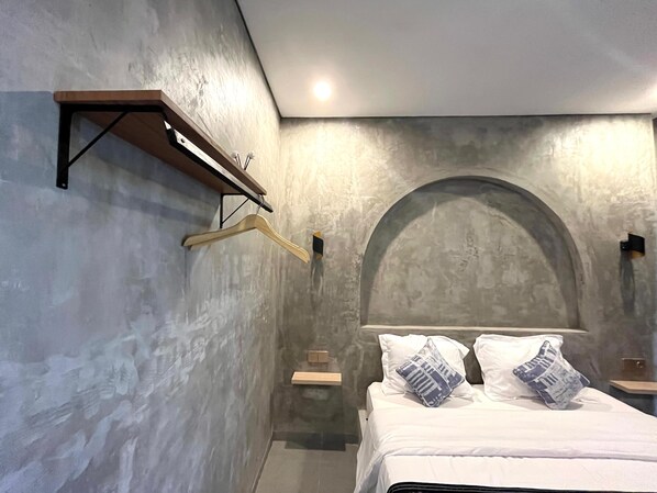 A bedroom with a bed, a wooden hanger, a shelf, and a wall-mounted light.