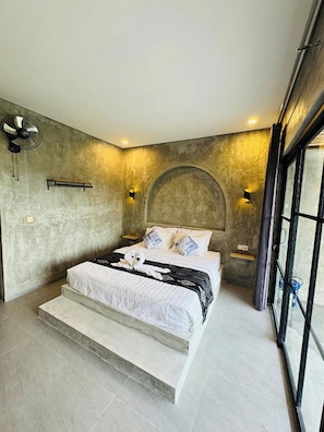 Une chambre moderne avec un grand lit, un ventilateur et une vue sur l’extérieur, visible à travers une porte vitrée.