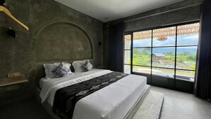 Een slaapkamer met een groot bed, uitzicht op groen en een betonnen muur.