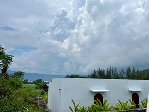A white building with arched windows and a flat roof, surrounded by greenery and a cloudy sky.