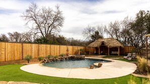 Un jardin avec une piscine, une clôture en bois, une structure à toit de chaume et une terrasse en pierre.
