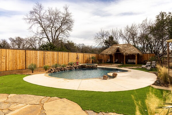 A backyard with a swimming pool, a wooden fence, a thatched-roof structure, and a stone patio.