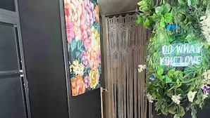Une pièce dotée d’un plancher en bois, d’un tableau floral aux couleurs vives accroché au mur, d’un rideau en macramé et d’une pancarte d’accueil agrémentée de verdure.