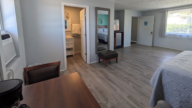 A spacious room with a wooden floor, a bed with a grey quilt, a small wooden table, and a chair. There is a bathroom with a white sink, a mirror, and a towel hanging on the wall. The room has a large window with curtains.