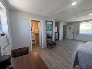 A spacious room with a wooden floor, a bed with a grey quilt, a small wooden table, and a chair. There is a bathroom with a white sink, a mirror, and a towel hanging on the wall. The room has a large window with curtains. - Lakeside Studio Apartment (Mooresburg)