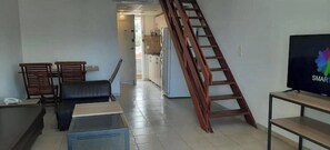 Una sala de estar con sofá, una mesa de centro, televisor y una escalera de madera que lleva a una zona de cocina.