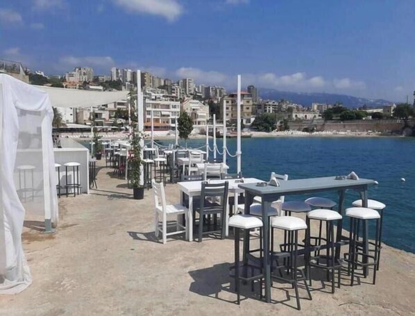 Un restaurante frente al mar con terraza, mesas y sillas blancas, y vistas al océano y edificios al fondo.