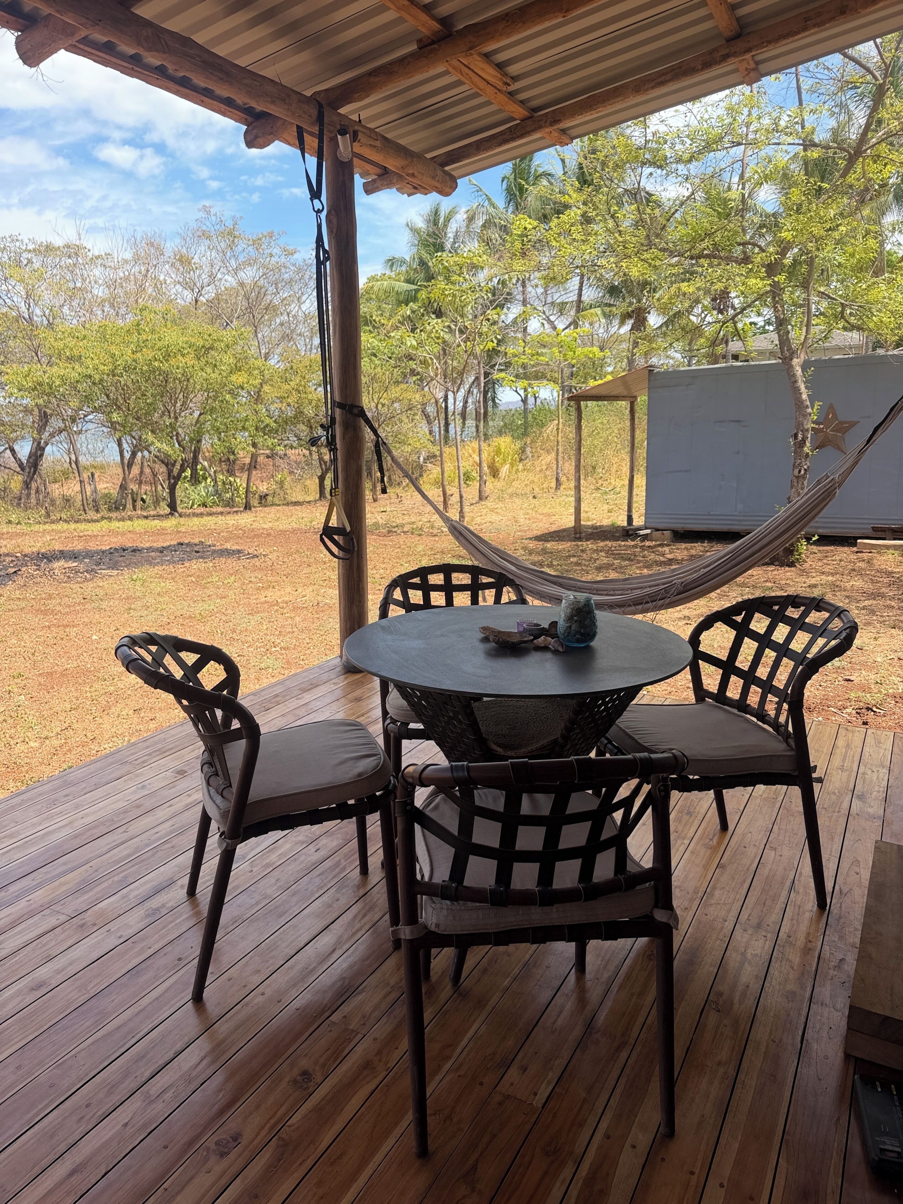 Eine Holzterrasse mit einem runden Tisch und vier Stühlen, einer Hängematte und einem überdachten Bereich mit Blick auf Bäume und ein Gebäude im Hintergrund.