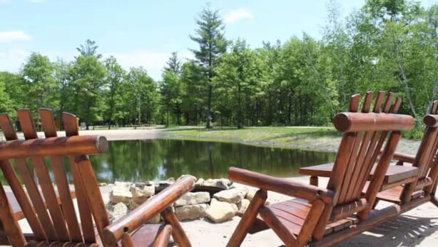 Eine Reihe aus Holz gefertigter Adirondack-Stühle, die auf einen stillen See mit umliegenden Bäumen ausgerichtet sind.