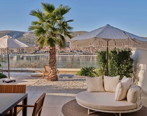 Un patio frente al mar con una palmera, un sofá redondo color beige con cojines, y vistas al océano y edificios a lo lejos.