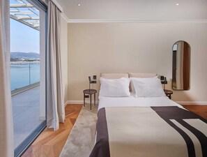 Habitación de hotel con una cama grande, un espejo redondo, mesitas de noche y vistas a una masa de agua desde un balcón.