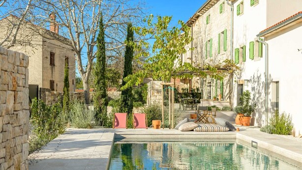 A pool area with a stone wall, a pergola with greenery, and a building with green shutters.