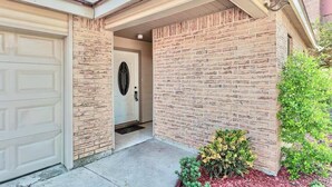 A residential entrance with a garage door, a brick wall, and a small garden with various plants. - Large  House in Peaceful McAllen. Large Pool, 10th/Trenton. Perfect Location (McAllen)