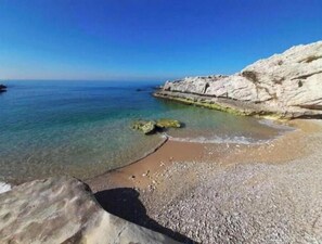 A coastal beach with clear water, rocky cliffs, and pebbly shore.