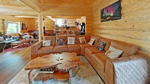 L’intérieur d’une cabane en rondins présente un grand plafond en bois, un ensemble de canapés en cuir, une table basse en bois et un tableau représentant une scène de forêt.