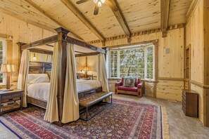 Una cama con dosel y cuatro columnas, un banco, un sofá rojo y un ventanal con vistas a árboles.