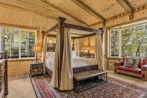 A four-poster bed with curtains, a bench, a red sofa, and a large window with a view of trees.