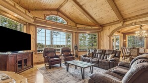 A spacious living room with wooden ceilings, large windows, leather furniture, and a flat-screen TV.
