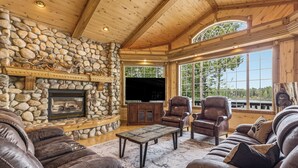 A stone fireplace, leather sofas, a wooden coffee table, and a TV in a cozy living room with large windows.
