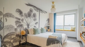 A bedroom with a large bed, a wall mural, a window with a view, and a bedside table with a lamp.