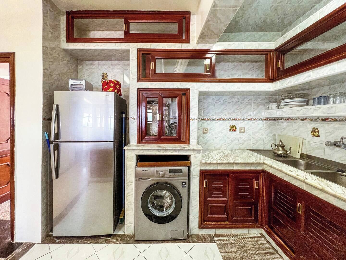 A compact kitchen with a stainless steel refrigerator, a washing machine, and wooden cabinets.