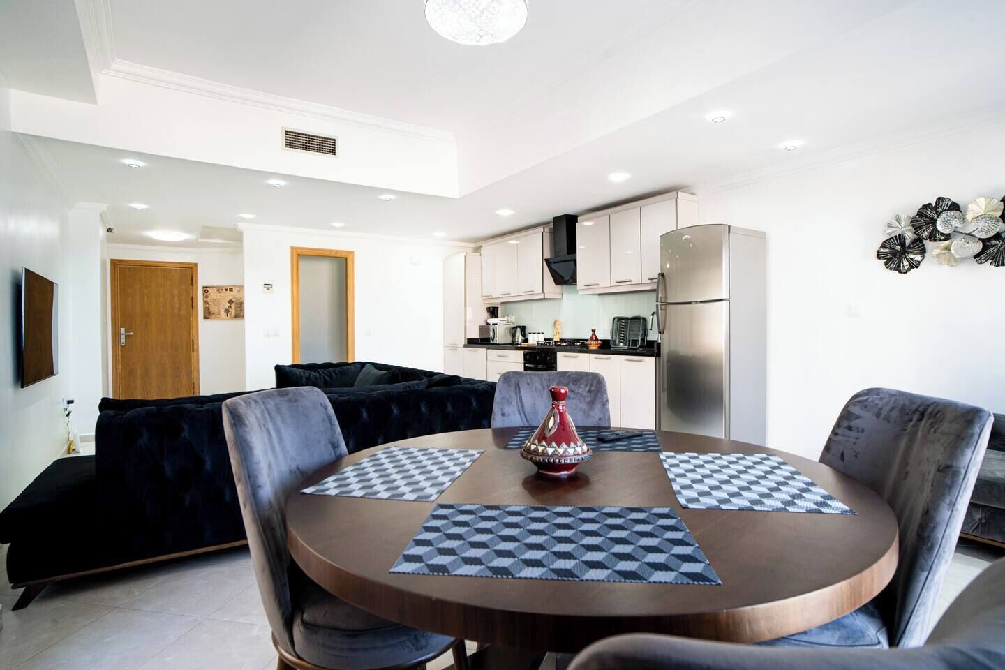 A modern living room with a round dining table, dark sofas, and a kitchen in the background.