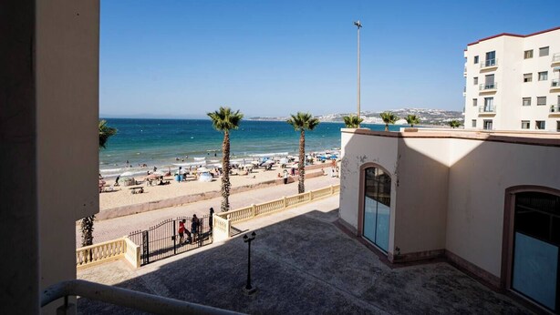 A balcony with a view of a beach, palm trees, and a clear blue sky. - The perfect coastal getaway, just steps from the beach, with a private balcony, open living area. (Tangier)